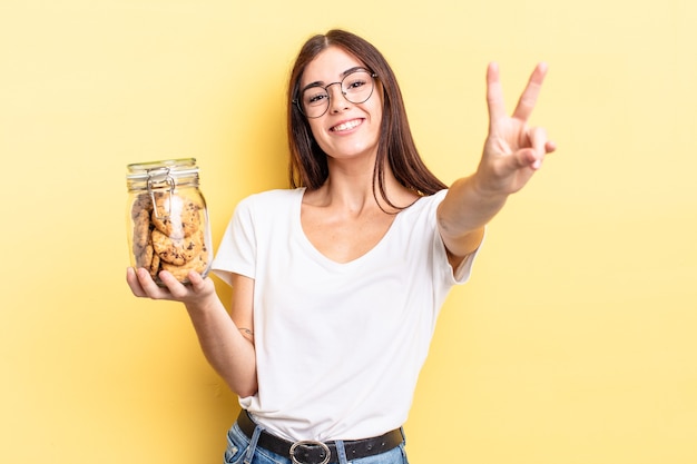 Young hispanic woman smiling and looking happy, gesturing victory or peace. cookies bottle concept