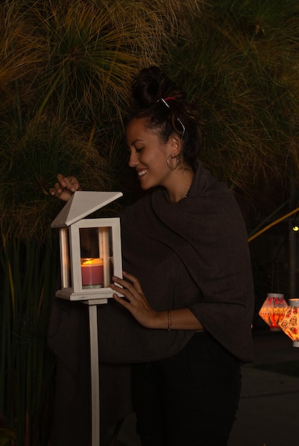 젊은 히스패닉계 여성이 촛불이 켜진 나무 랜턴의 뚜껑을 열고 웃고 있습니다.