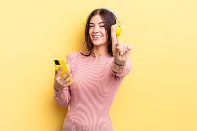 Giovane donna ispanica che sorride con orgoglio e sicurezza facendo il numero uno. concetto di cuffie e telefono