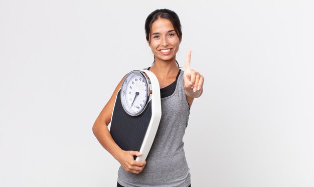 Young hispanic woman smiling proudly and confidently making number one and holding a weight scale. diet concept