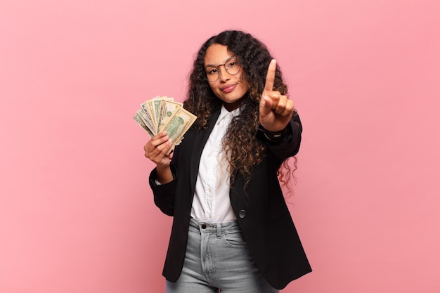 Young hispanic woman smiling proudly and confidently making number one pose triumphantly, feeling like a leader. dollar banknotes concept