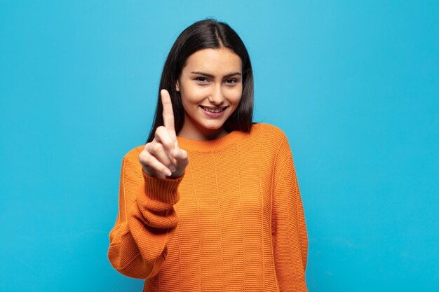 Young hispanic woman smiling proudly and confidently making number one pose triumphantly, feeling like a leader