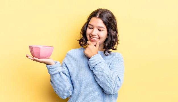 Young hispanic woman smiling with a happy, confident expression with hand on chin. empty bowl concept