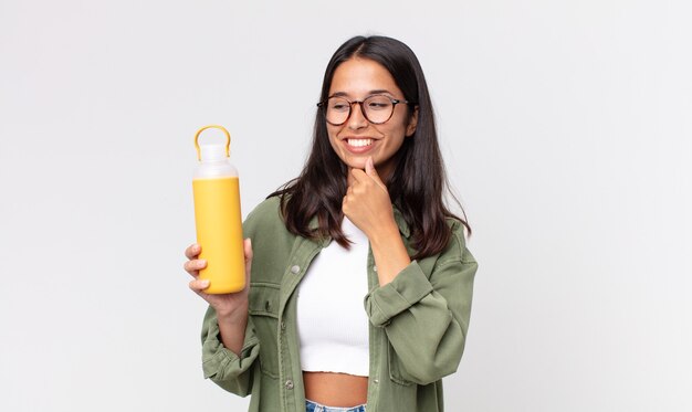 Young hispanic woman smiling with a happy, confident expression with hand on chin and holding a coffee thermos