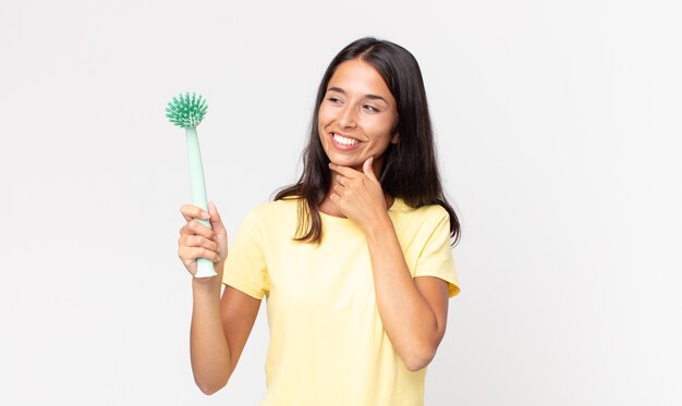 Young hispanic woman smiling with a happy, confident expression with hand on chin and holding a dish cleaning brush