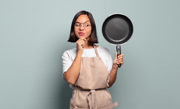 ヒスパニック系の若い女性が、あごに手を当てて幸せで自信に満ちた表情で笑って、不思議に思って横を向いています