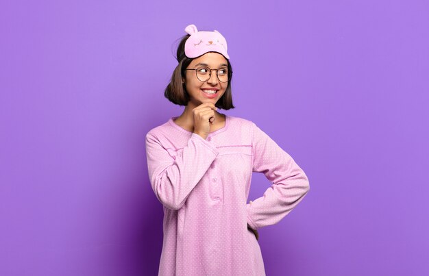 Young hispanic woman smiling with a happy, confident expression with hand on chin, wondering and looking to the side