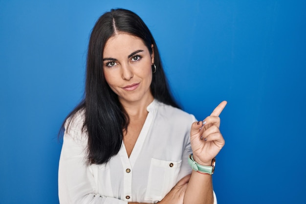 Young hispanic woman standing over blue background pointing with hand finger to the side showing advertisement serious and calm face
