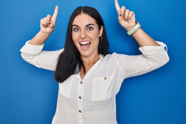 Young hispanic woman standing over blue background smiling amazed and surprised and pointing up with fingers and raised arms.