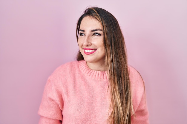 Young hispanic woman standing over pink background looking away to side with smile on face, natural expression. laughing confident.