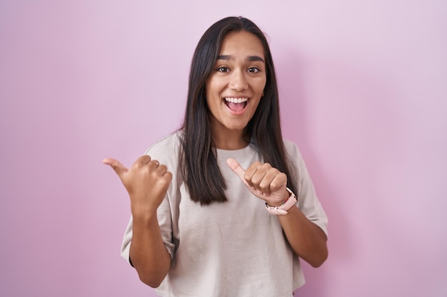 Young hispanic woman standing over pink background pointing to the back behind with hand and thumbs up smiling confident