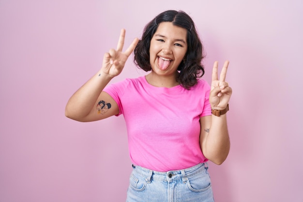 Young hispanic woman standing over pink background smiling with tongue out showing fingers of both hands doing victory sign number two