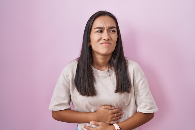 Young hispanic woman standing over pink background with hand on stomach because indigestion, painful illness feeling unwell. ache concept.