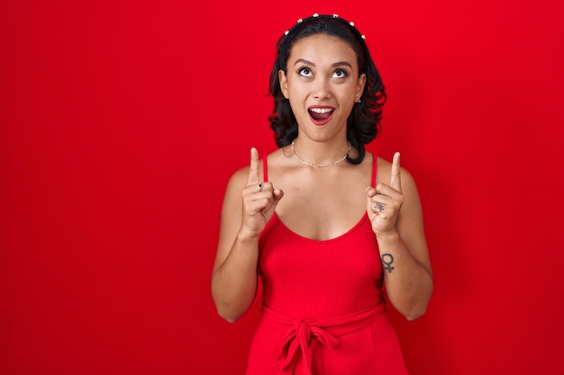 Young hispanic woman standing over red background amazed and surprised looking up and pointing with fingers and raised arms.