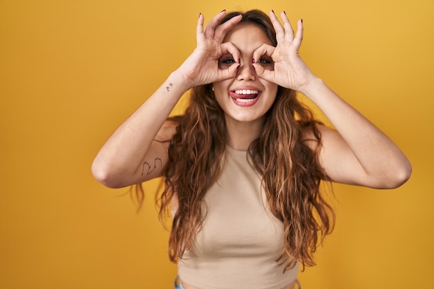 Young hispanic woman standing over yellow background doing ok gesture like binoculars sticking tongue out eyes looking through fingers crazy expression