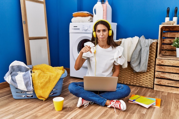 Young hispanic woman studying while waiting for laundry pointing down looking sad and upset indicating direction with fingers unhappy and depressed