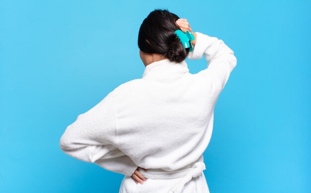 Young hispanic woman thinking or doubting, scratching head, feeling puzzled and confused, back or rear view. bathrobe concept