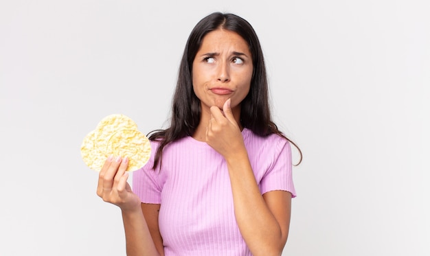 Young hispanic woman thinking, feeling doubtful and confused and holding a rice cookie. diet concept