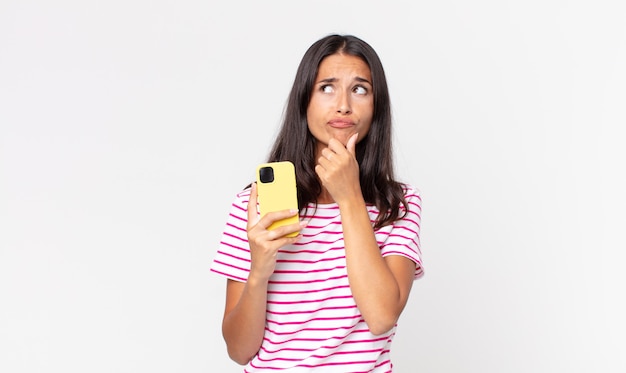 young hispanic woman thinking, feeling doubtful and confused and holding a smartphone