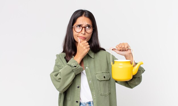 Young hispanic woman thinking, feeling doubtful and confused and holding a tea pot