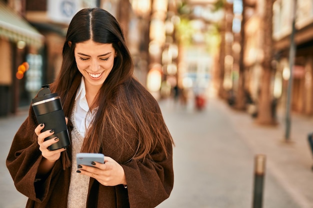 Young hispanic woman using smartphone and drinking coffee at the city