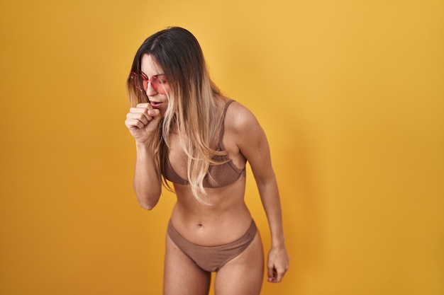 Young hispanic woman wearing bikini over yellow background feeling unwell and coughing as symptom for cold or bronchitis health care concept