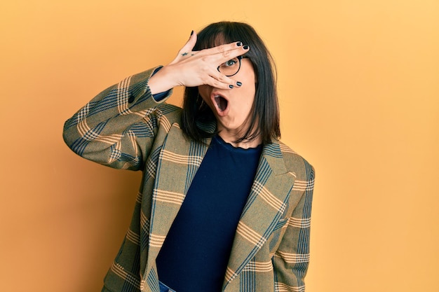 Young hispanic woman wearing business style and glasses peeking in shock covering face and eyes with hand looking through fingers afraid