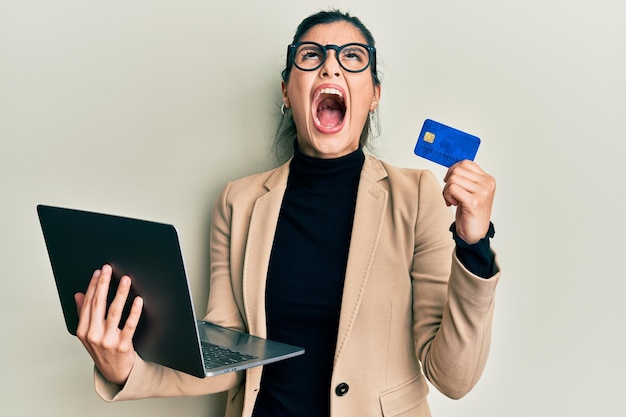 Young hispanic woman wearing business style holding laptop and credit card angry and mad screaming frustrated and furious shouting with anger looking up