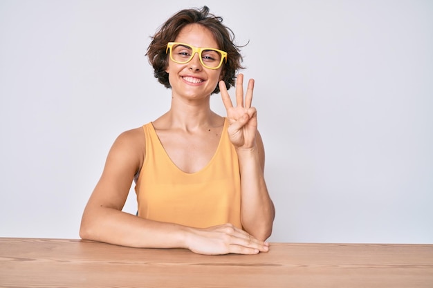 Young hispanic woman wearing casual clothes and glasses sitting on the table showing and pointing up with fingers number three while smiling confident and happy.