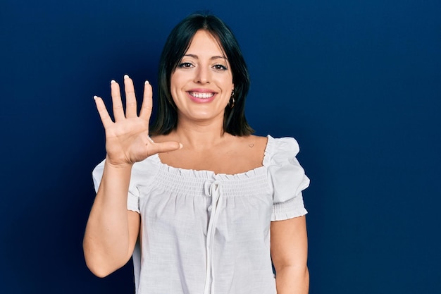 カジュアルな服を着た若いヒスパニック系の女性が、自信を持って幸せそうに微笑みながら、5番の指で上向きに指差しています。