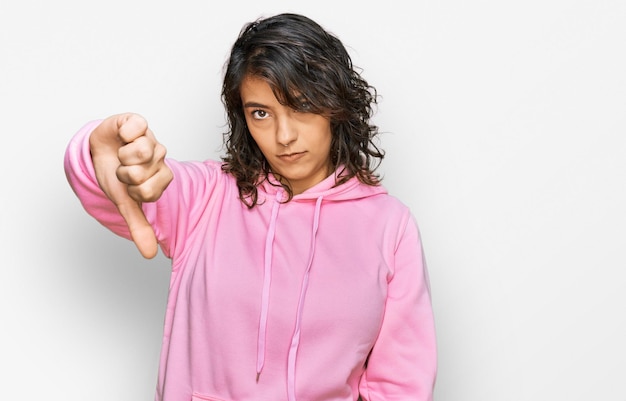 カジュアルなスウェットシャツを着たヒスパニック系の若い女性が不幸で怒っているように見え、親指を下げるジェスチャーで悪い表情で拒絶と否定性を示す