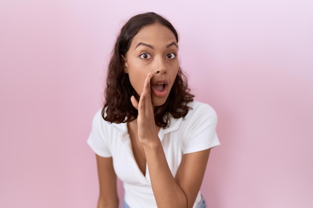 Young hispanic woman wearing casual white t shirt hand on mouth telling secret rumor, whispering malicious talk conversation