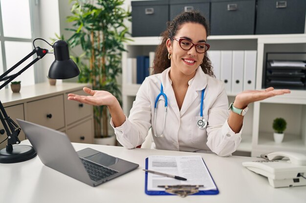 Young hispanic woman wearing doctor uniform and stethoscope smiling showing both hands open palms presenting and advertising comparison and balance