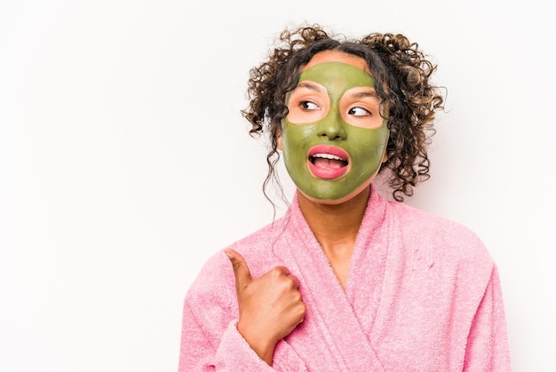 Young hispanic woman wearing a facial mask isolated on white background points with thumb finger away laughing and carefree