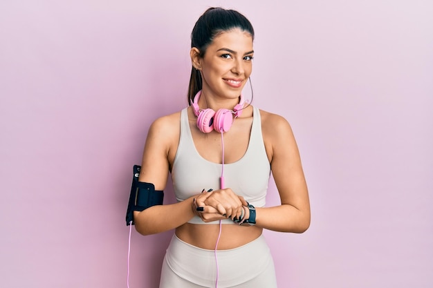 Young hispanic woman wearing gym clothes and using headphones with hands together and crossed fingers smiling relaxed and cheerful. success and optimistic