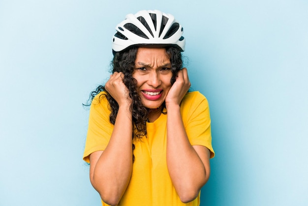 Young hispanic woman wearing a helmet bike isolated on blue background covering ears with hands