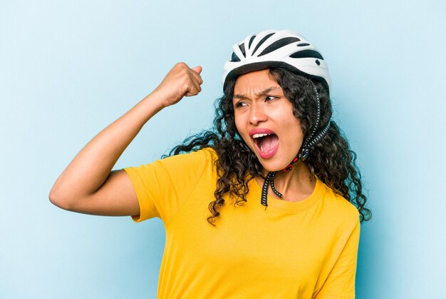 Young hispanic woman wearing a helmet bike isolated on blue background raising fist after a victory winner concept