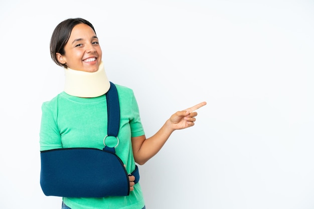 Young hispanic woman wearing a neck brace and sling isolated on white background pointing finger to the side