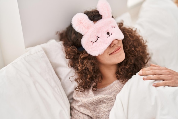 Young hispanic woman wearing sleepmask sleeping on bed at bedroom