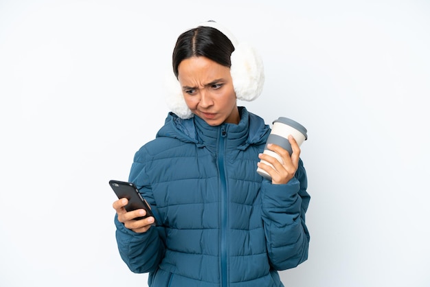 Young hispanic woman wearing a winter earmuffs isolated on white background holding coffee to take away and a mobile