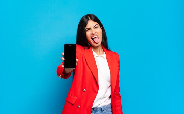 Young hispanic woman with cheerful, carefree, rebellious attitude, joking and sticking tongue out, having fun, phone screen copy space