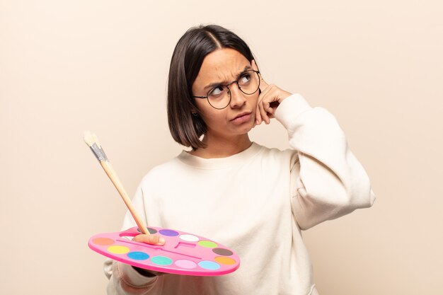 Young hispanic woman with a concentrated look, wondering with a doubtful expression, looking up and to the side