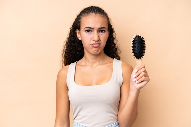 Photo young hispanic woman with hair comb isolated on beige background with sad expression