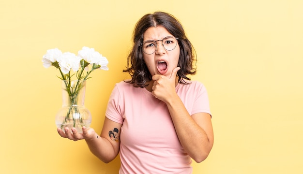 Young hispanic woman with mouth and eyes wide open and hand on chin. flowers pot concept