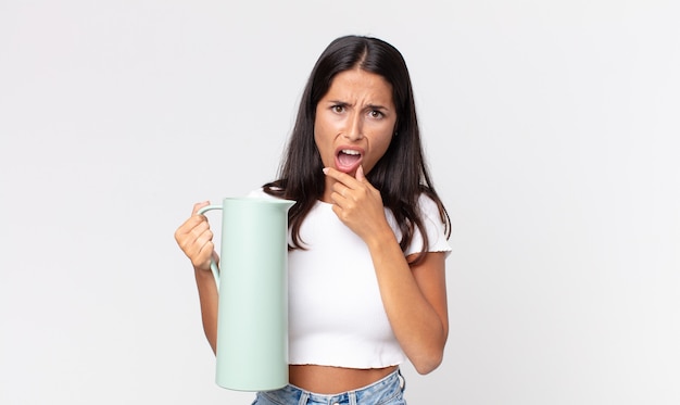 Young hispanic woman with mouth and eyes wide open and hand on chin and holding a coffee thermos