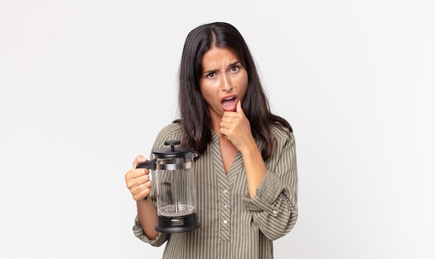 Young hispanic woman with mouth and eyes wide open and hand on chin and holding a manual coffee maker