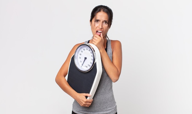 Young hispanic woman with mouth and eyes wide open and hand on chin and holding a weight scale. diet concept