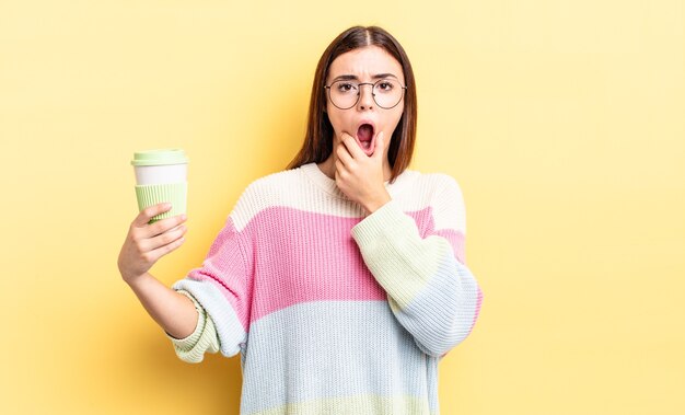 Young hispanic woman with mouth and eyes wide open and hand on chin. take away coffee concept