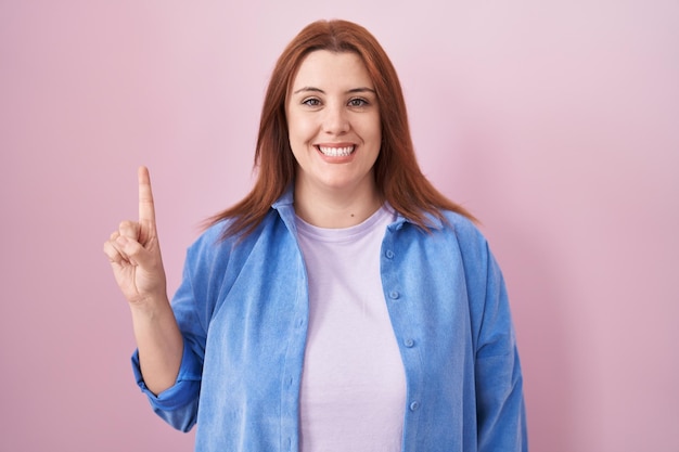 Young hispanic woman with red hair standing over pink background showing and pointing up with finger number one while smiling confident and happy.