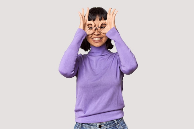 Молодая латиноамериканка с короткими черными волосами в студии показывает знак "окей" над глазами